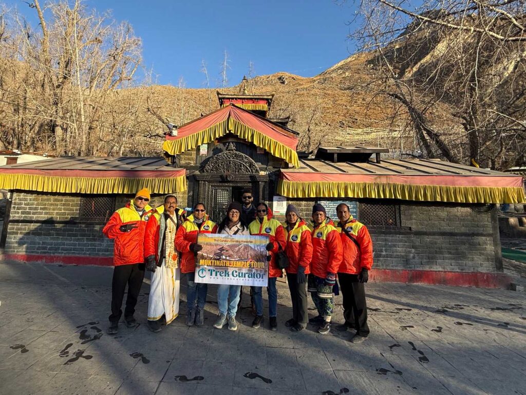 A group of trekkers wearing matching orange and yellow jackets pose in front of the Muktinath Temple in the mountains, holding a Trek Curator banner on a sunny day.