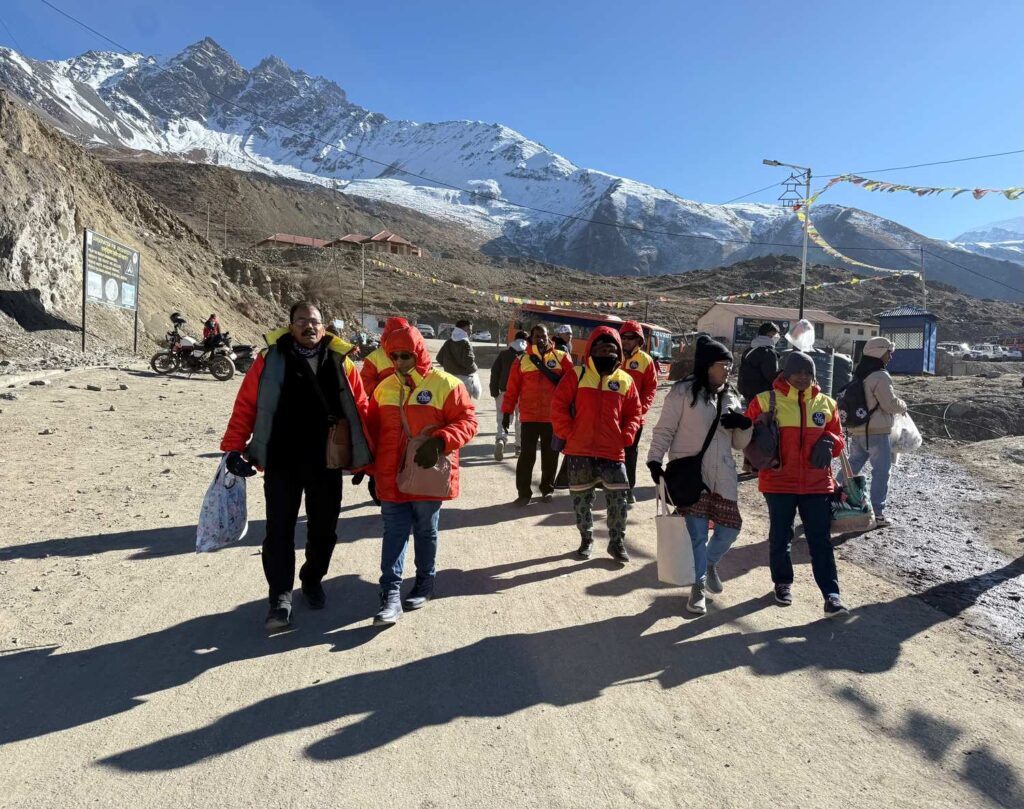 A group of travelers walking along a mountain road with snow covered peaks and prayer flags in the background under a clear blue sky.