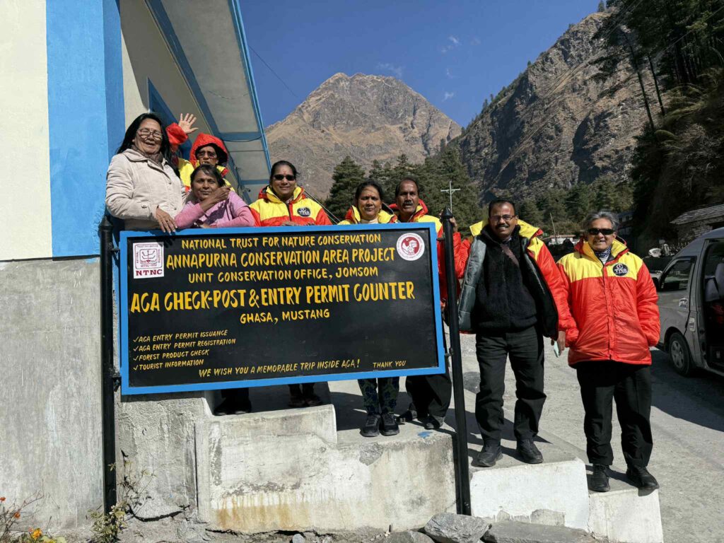 Group of travelers posing at the ACA check-post and entry permit counter in Ghansa, Mustang, during an Upper Mustang jeep tour, with rugged Himalayan mountains and a bright blue sky in the background.