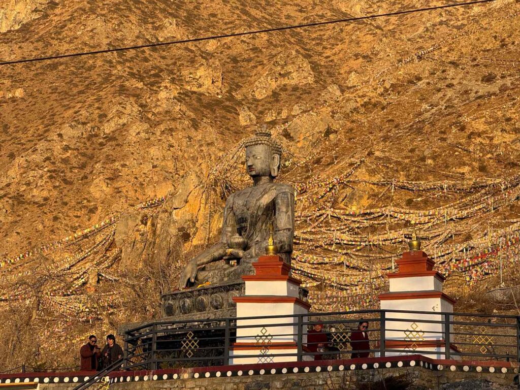 Large seated Buddha statue on a mountainside, surrounded by colorful prayer flags, with a few visitors standing on a terrace below.